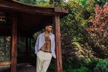 young african american man standing with hands in pocket of white trousers in wooden gazebo in park