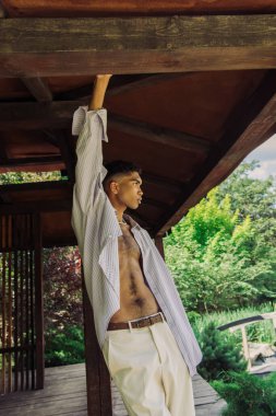 young african american man in striped shirt looking away while standing in wooden gazebo with raised hand