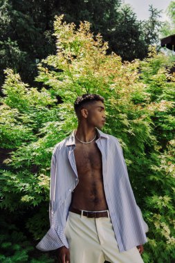 young african american man in striped shirt standing near green bush in park