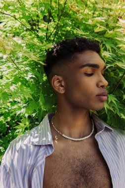 portrait of pierced african american man in beads and blue shirt near bush with green leaves