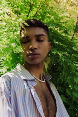 portrait of african american man with piercing near bush with green leaves