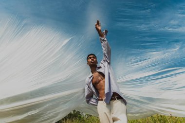 stylish african american man with closed eyes and raised hand under cellophane film outdoors