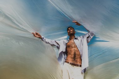 trendy african american man with closed eyes posing under polyethylene outdoors