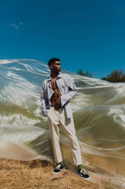young african american man with hand in pocket of white trousers standing in meadow near polyethylene film