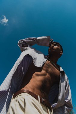 low angle view of trendy african american man in shirt posing against blue sky