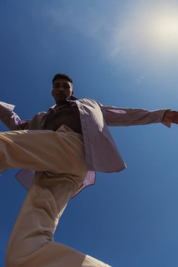 bottom view of african american man in trendy clothes under blue and clear sky