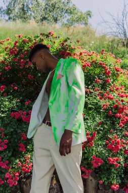 Afro-Amerikalı trend yeşil beyaz ceketli adam parktaki filizlenen çalılara bakıyor.