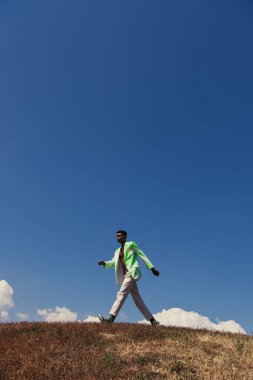 side view of african american man in blazer and pants walking in field under clear sky clipart