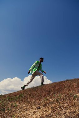side view of stylish african american man walking on hill against sky with white clouds