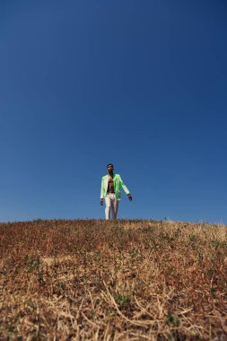 trendy african american man walking in meadow under blue and clear sky 