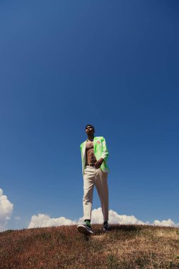 full length of african american man in trendy clothes walking in meadow under blue sky