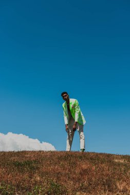 african american man in fashionable outfit standing in field under blue sky on summer day