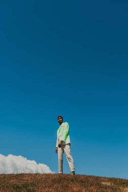 african american man in stylish blazer and trousers standing in meadow under blue sky