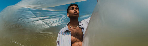 african american man in shirt and necklace standing with closed eyes under cellophane film, banner