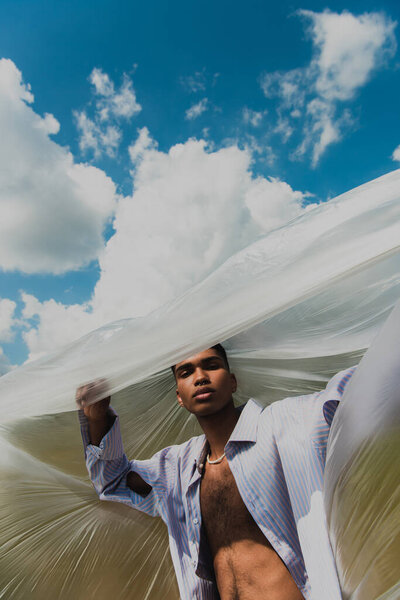 african american man in white shirt looking at camera under cellophane film and cloudy sky