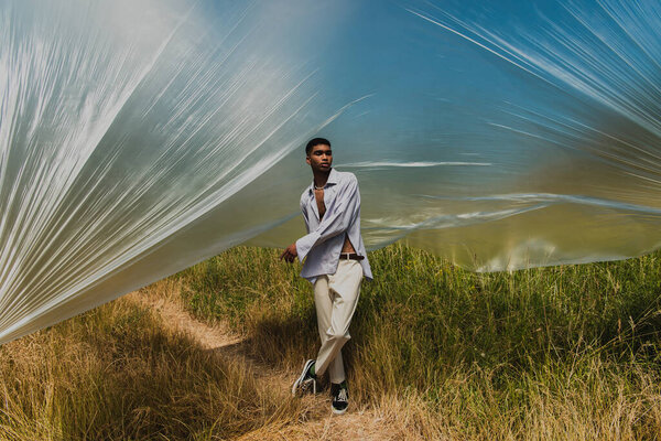 full length of african american man in summer outfit standing under polyethylene film in grassy meadow