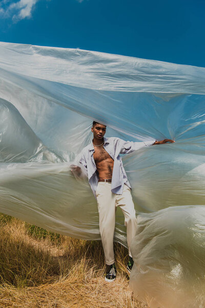 african american man in trendy shirt and trousers standing near cellophane film in field
