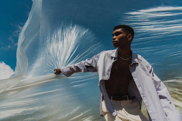 low angle view of trendy african american man under cellophane film and blue sky