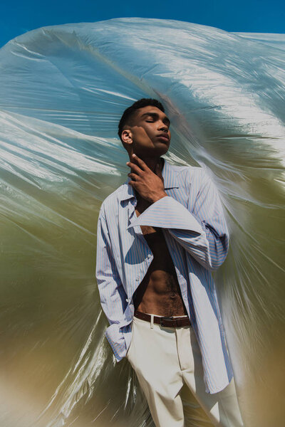 fashionable african american man touching neck while standing with hand in pocket near cellophane film