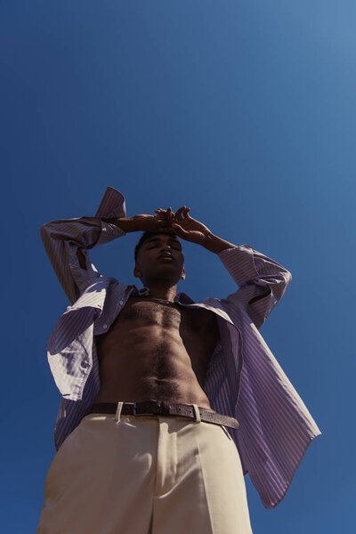 low angle view of african american man in shirt posing with hands near head under blue sky