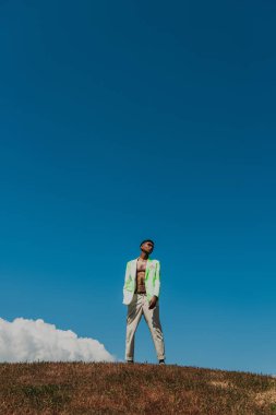 fashionable african american man in summer clothes standing in field under blue sky