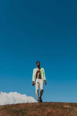 full length of stylish african american man in summer outfit walking in meadow under clear sky