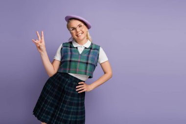 Smiling student in eyeglasses and beret showing peace gesture isolated on purple 