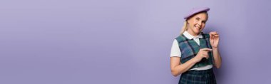 Happy blonde student in beret holding eyeglasses on purple background, banner  
