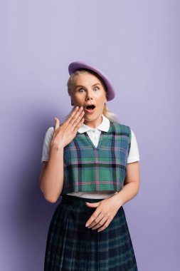 Shocked student in beret looking at camera on purple background 