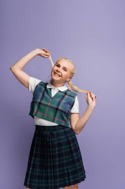 Positive student in plaid uniform holding blonde hair isolated on purple 