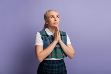 Student in checkered uniform doing praying hands isolated on purple 