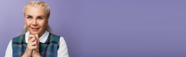 Blonde student with clenched hands smiling at camera isolated on purple, banner 