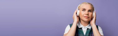 Student in shirt and vest listening music in headphones isolated on purple, banner 