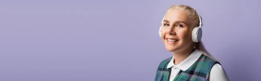 Positive blonde student in headphones looking at camera isolated on purple, banner 