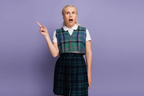 Shocked blonde student pointing with finger on purple background