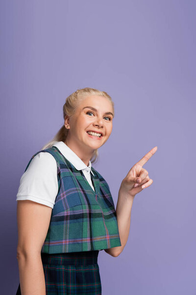Positive blonde student pointing with finger and looking at camera isolated on purple 