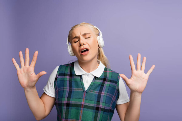 Blonde student in headphones singing isolated on purple 