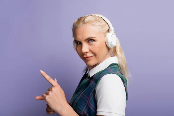 Blonde student in headphones pointing with finger isolated on purple