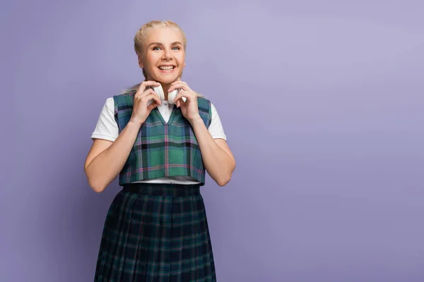 Happy student in uniform holding headphones isolated on purple