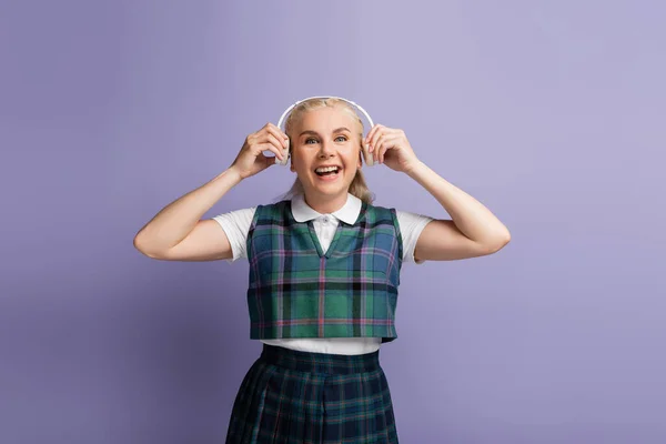 Positive student in uniform holding headphones near ears isolated on purple