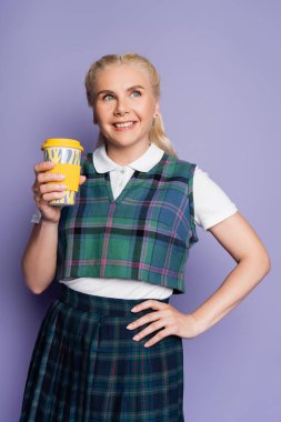 Cheerful student in checkered uniform holding coffee to go on purple background 