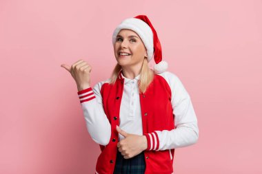 Smiling woman in santa hat pointing with finger isolated on pink 