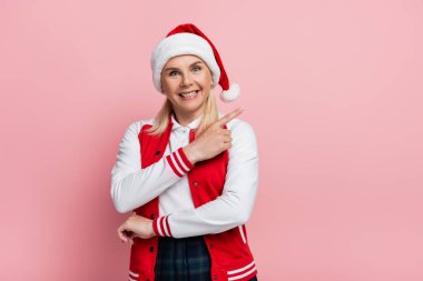 Positive woman in santa hat pointing with finger and looking at camera isolated on pink 