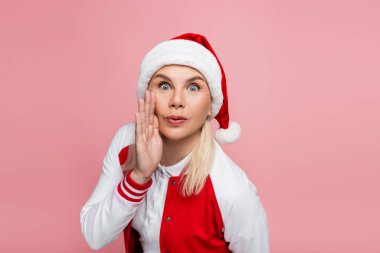 Astonished woman in santa hat holding hand near cheek isolated on pink 