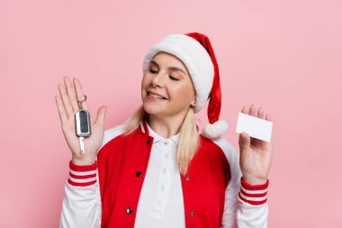 Smiling blonde woman in santa hat holding car key and empty driving license isolated on pink 
