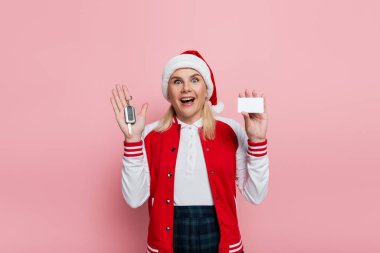 Excited woman in santa hat holding car key and empty driving license isolated on pink 