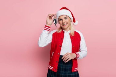 Positive woman in santa hat holding car key isolated on pink 