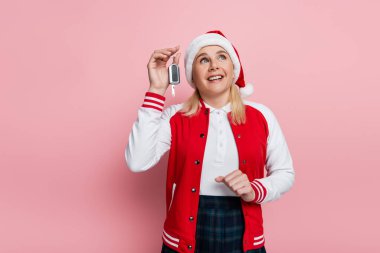 Positive woman in red santa hat holding car key and looking up isolated on pink 