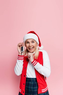 Excited woman in santa hat holding car key and showing yes gesture isolated on pink 