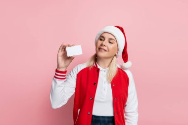 Blonde woman in red santa hat holding empty driving license isolated on pink 